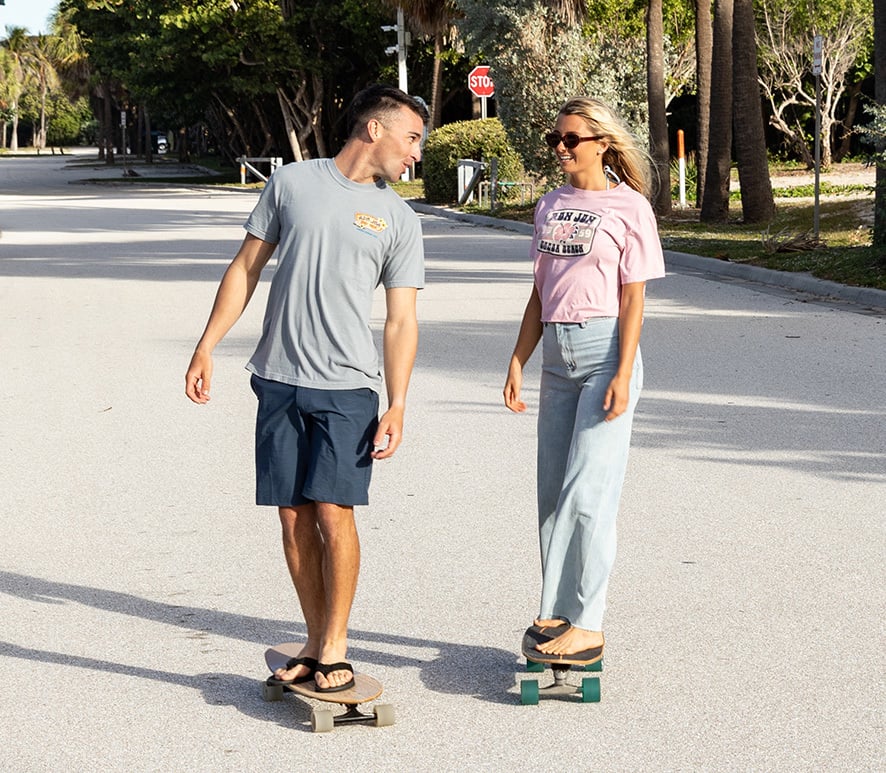A man and a woman ride skateboards on a sunny street, looking at each other and smiling.