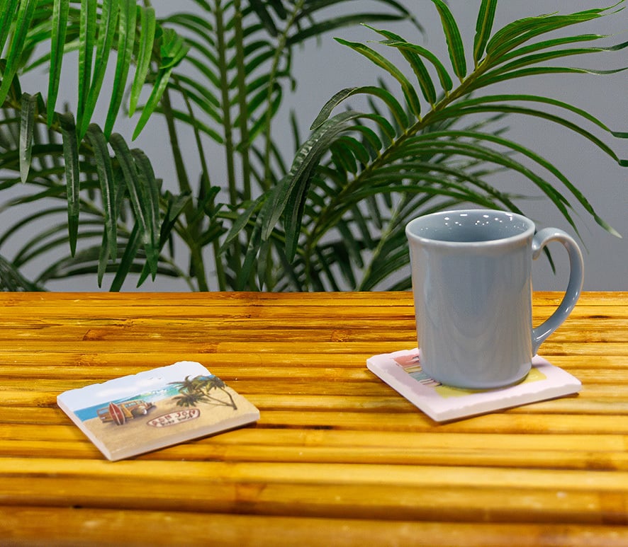 A gray ceramic mug sits on a coaster on a bamboo table, with another coaster nearby. Green tropical plants are in the background.