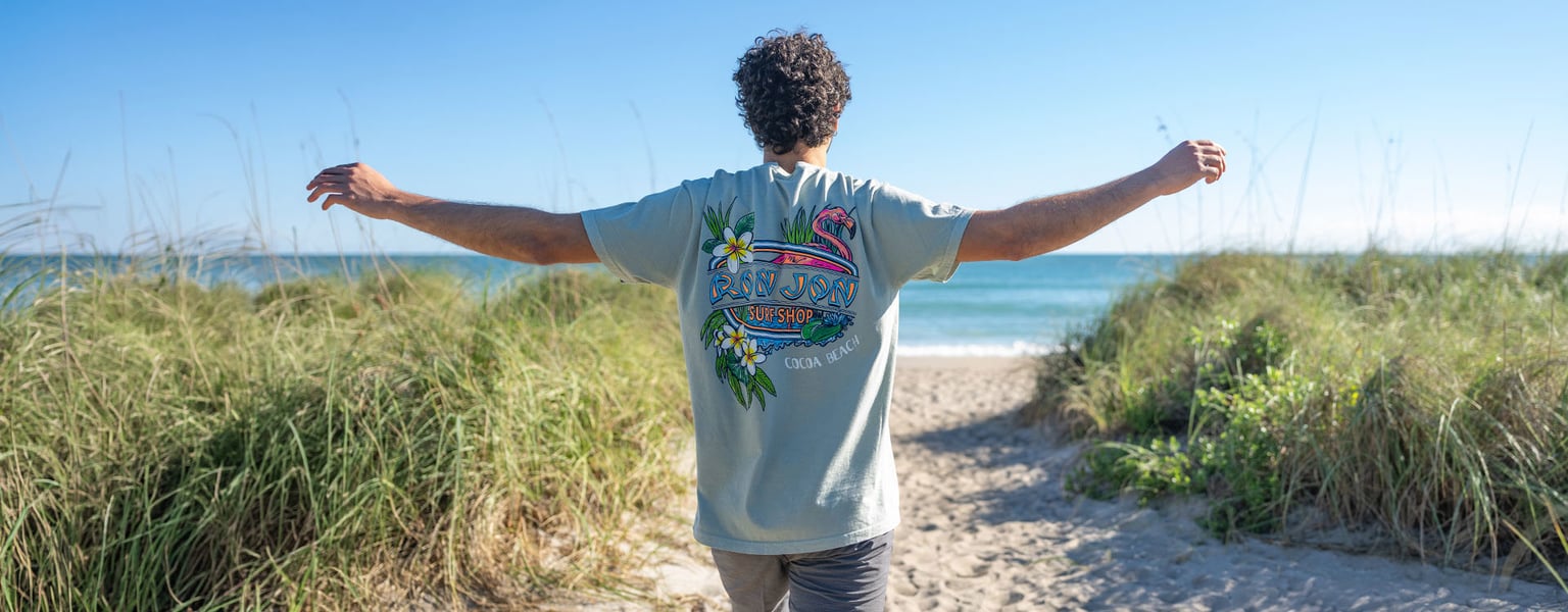 A person with curly hair stands on a sandy path between tall grass, arms outstretched, facing the ocean under a clear sky.