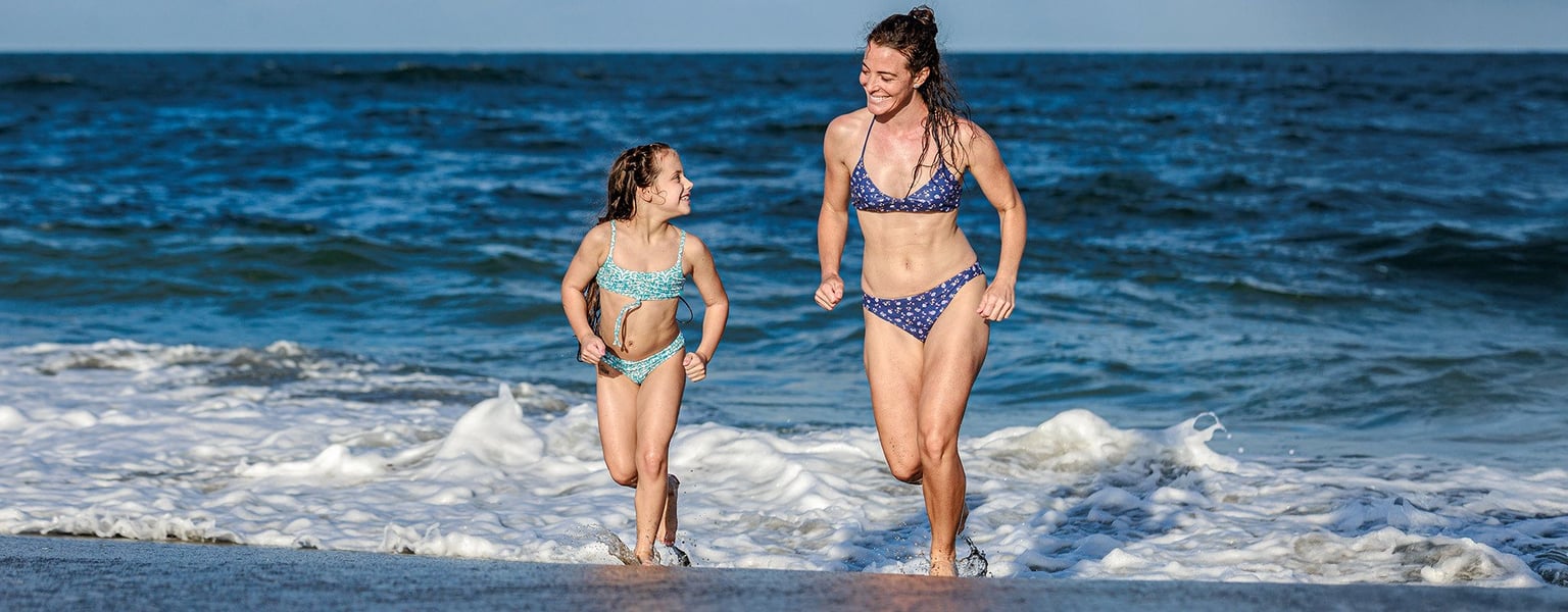 mother and daughter running at the water's edge wearing two-piece swimwear sets