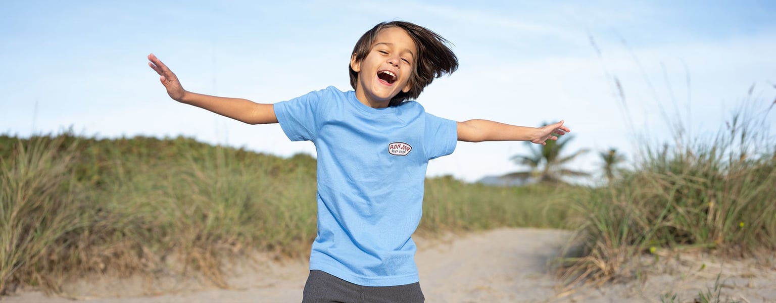 A young child wearing a blue t-shirt stands with arms outstretched and mouth open, outdoors on a sandy path with grass and a palm tree in the background.