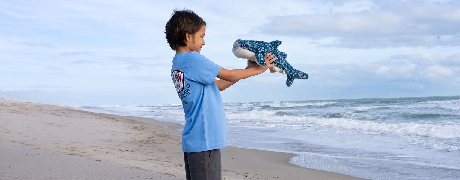 A young child in a blue shirt stands on a sandy beach, holding up a plush whale shark toy near the ocean waves under a partly cloudy sky.