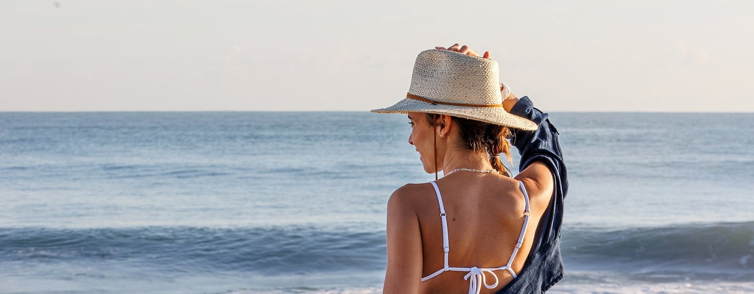 woman in swimwear standing at the oceans edge with a classic straw hat on