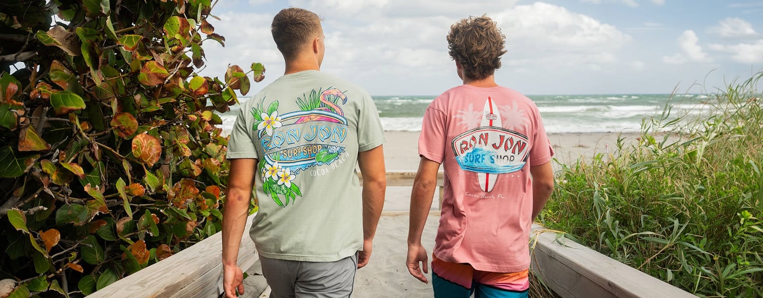 Two men wearing colorful graphic t-shirts walk on a wooden path toward a sandy beach with ocean waves in the background.