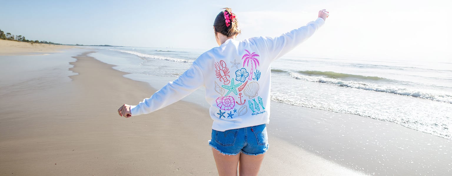 A person wearing a white sweatshirt with a colorful beach-themed graphic print stands on a sandy beach with their arms outstretched.