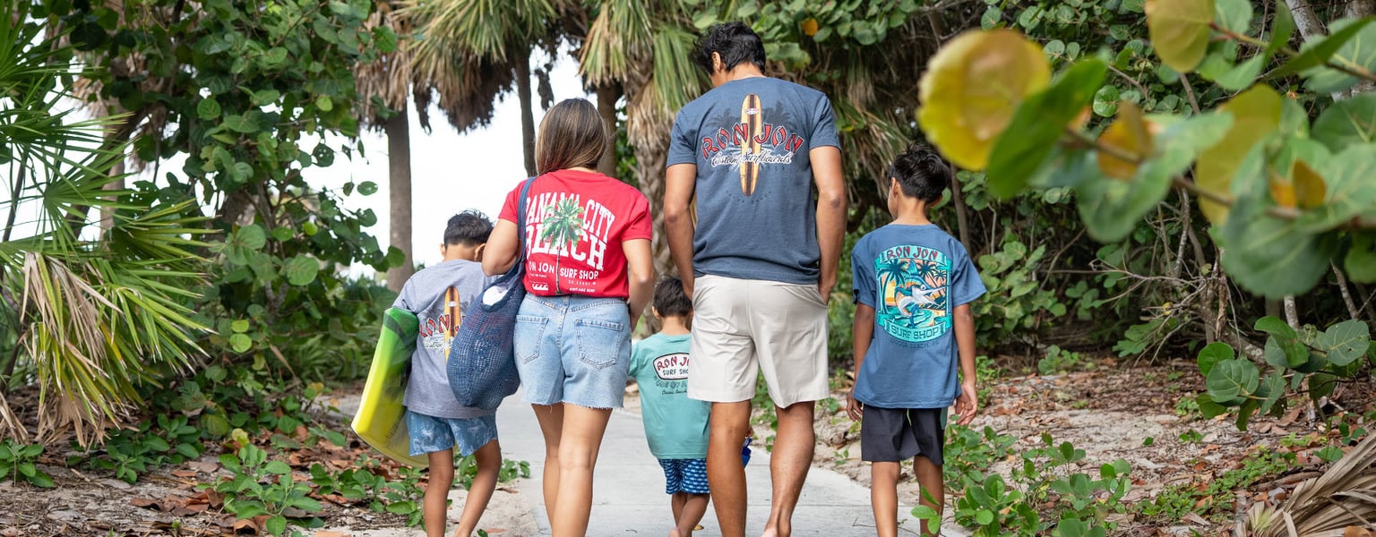 A family of five walks on a sandy path surrounded by greenery, all wearing colorful T-shirts and shorts, heading toward the beach.