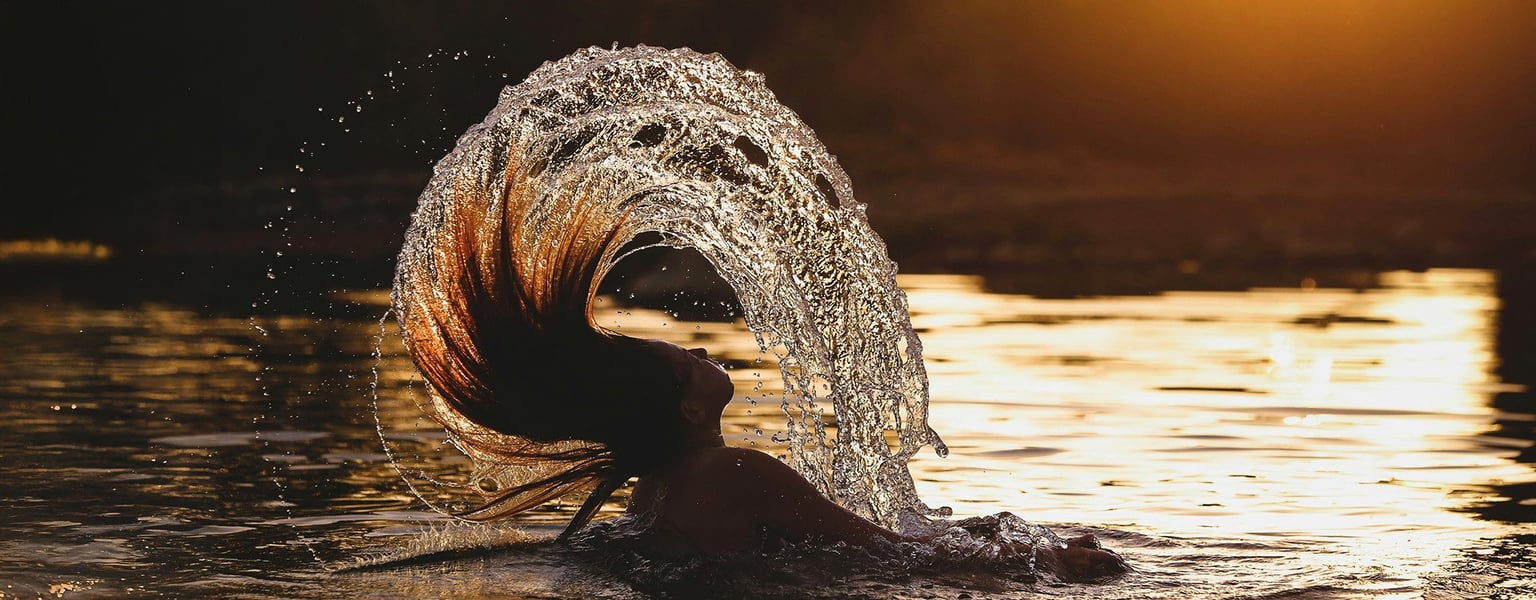 a person in a body of water flipping their hair, creating an arc of water at sunset