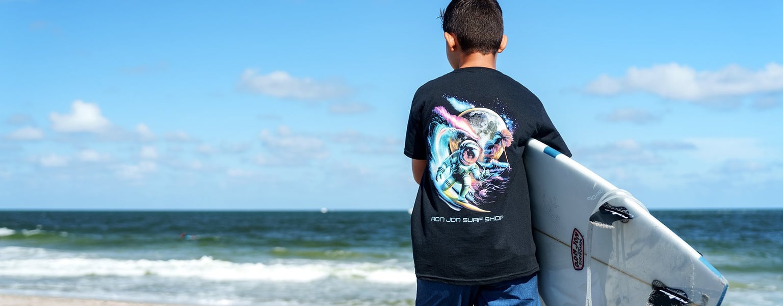 young person carrying a surfboard on the beach while wearing a space themed ron jon tee shirt