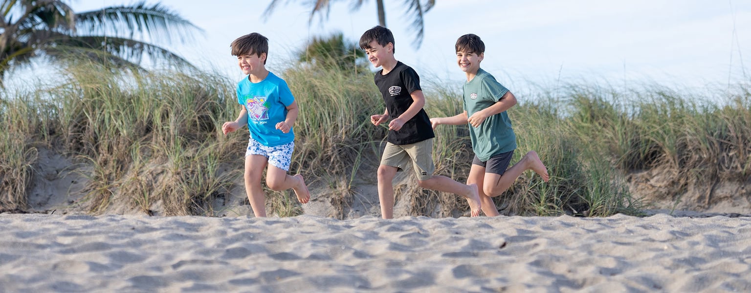 Three young boys running along a sandy beach with dune grass in the background.