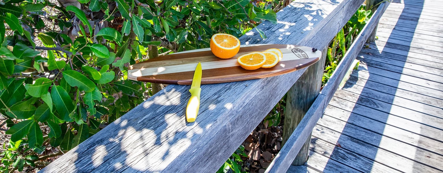 A wooden surfboard shaped cutting board with a sliced orange and a yellow knife sits on a wooden railing next to green foliage and a boardwalk.