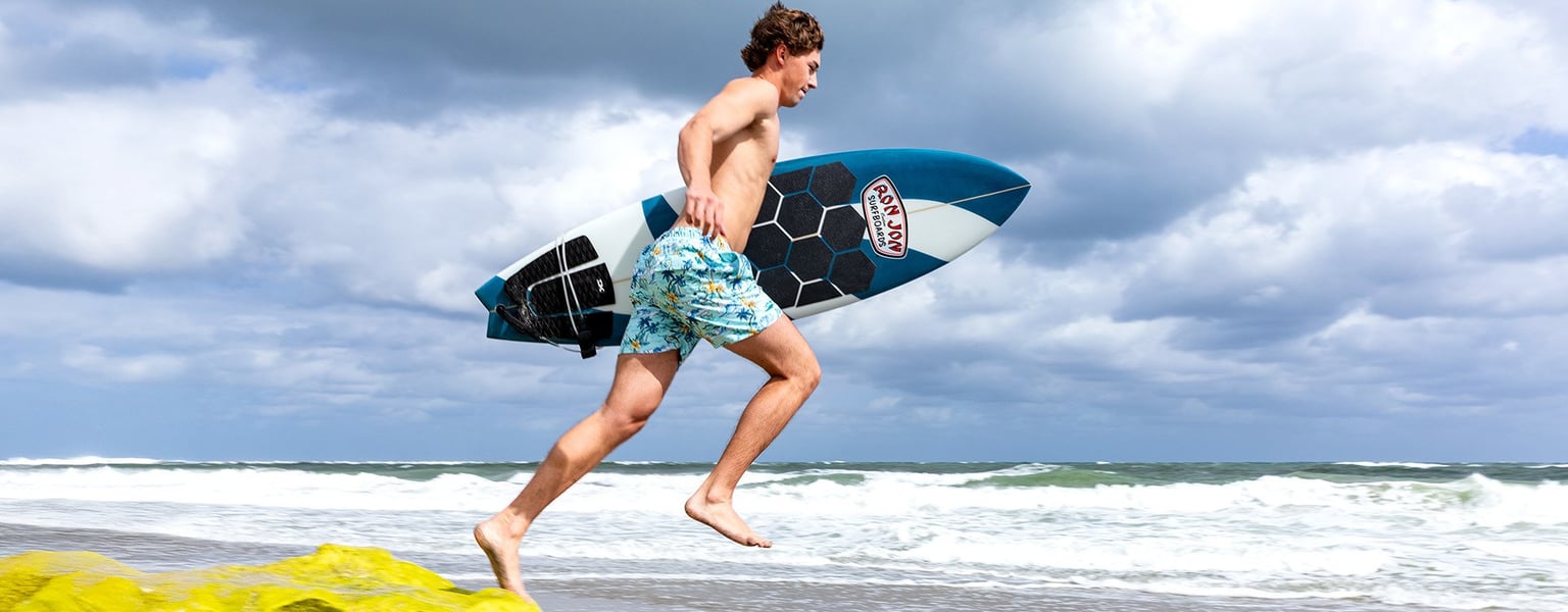 guy leaping toward the ocean wearing swimsuit and carrying a surfboard