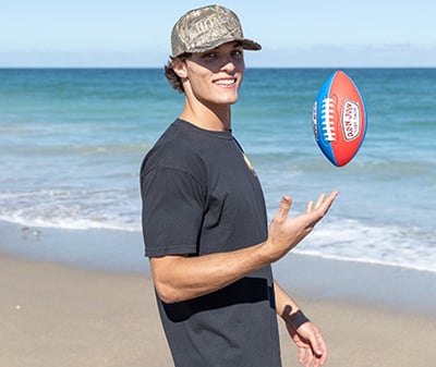 A man in a black shirt and camo hat stands on a beach, tossing a red and blue Ron Jon football in the air, with the ocean and sky in the background.