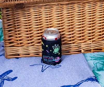 A soda can with a black alien-themed sleeve sits on a blue towel with starfish designs, in front of a woven picnic basket.