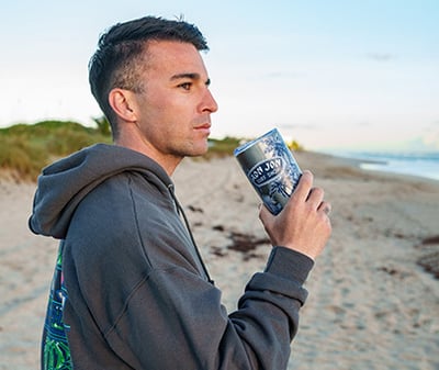 A man wearing a dark hoodie stands on a sandy beach, holding a steel tumbler and looking into the distance.