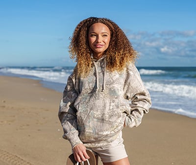 A woman with curly hair wearing a camouflage hoodie and tan shorts stands on a sandy beach with waves and blue sky in the background.