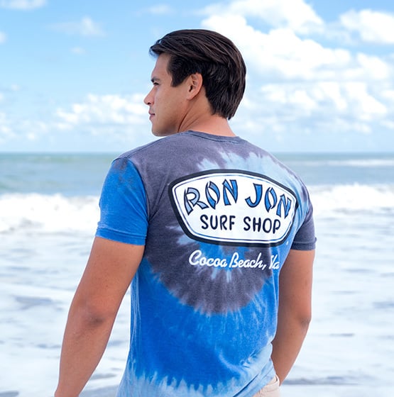 man standing on the beach looking out to the ocean while wearing a ron jon surf shop tie-dye t-shirt