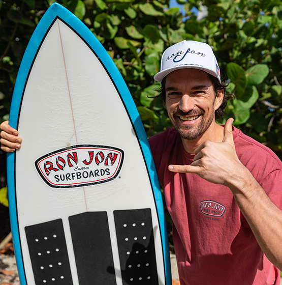 Man in a red Ron Jon Surf Shop shirt and white hat holds a Ron Jon surfboard and makes a shaka hand gesture, standing outdoors with green foliage in the background.