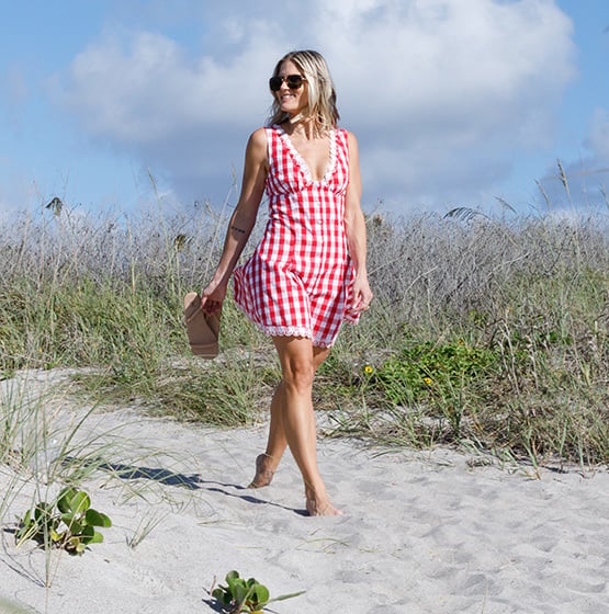 Woman in a red and white checkered sleeveless dress walks barefoot on a sandy beach, holding sandals, with grass and blue sky in the background.