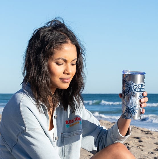 Woman sitting on a sandy beach holding a Ron Jon Surf Shop tumbler, with the ocean and sky in the background.